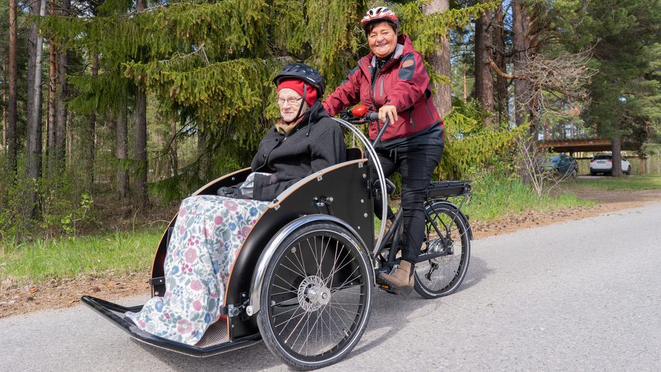 Kvinna som jobbar inom äldreomsorgen skjutsar äldre kvinna på en parcykel.