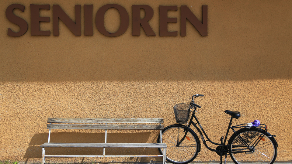 Solbelyst rappad husfasad med träbänk och parkerad damcykel framför. På väggen står Senioren.