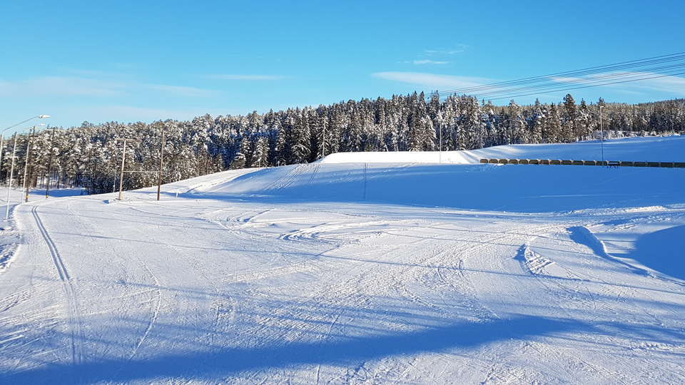 Snötäckt yta med en skidskyttevall i bakgrunden. Klarblå himmel och granskog i fonden.