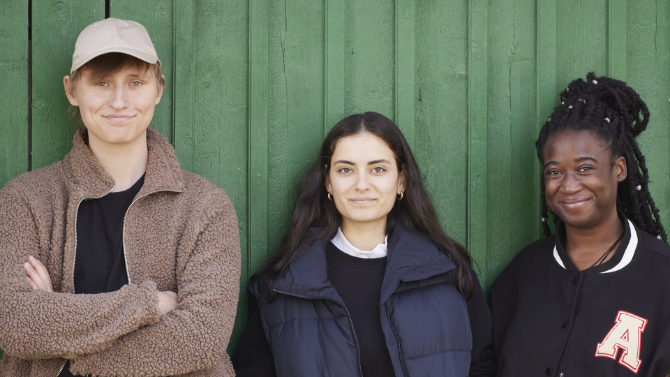 Tre ungdomar står lutade mot en grön vägg