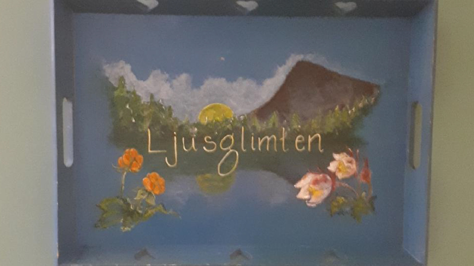 Bild av en bricka med målad bild av en fjälltopp, mosippa, hjortronblom och en sol.