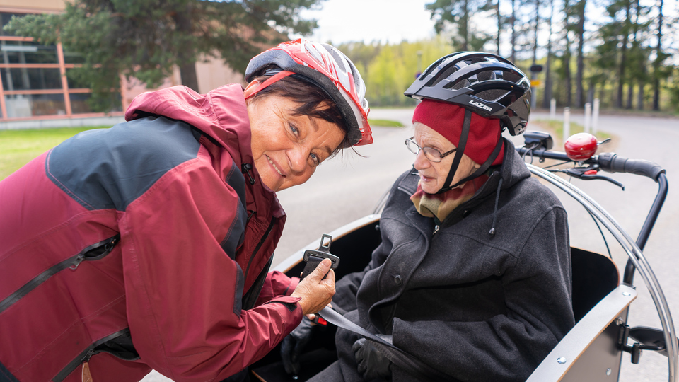 Kvinna som jobbar inom äldreomsorgen skjutsar äldre kvinna på en parcykel.