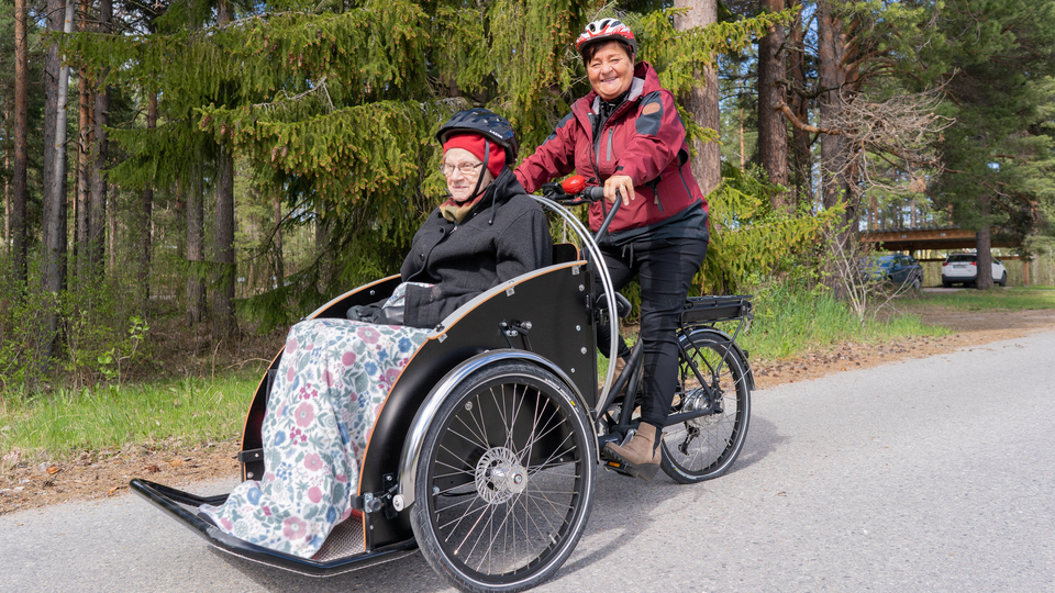 Kvinna som jobbar inom äldreomsorgen skjutsar äldre kvinna på en parcykel.
