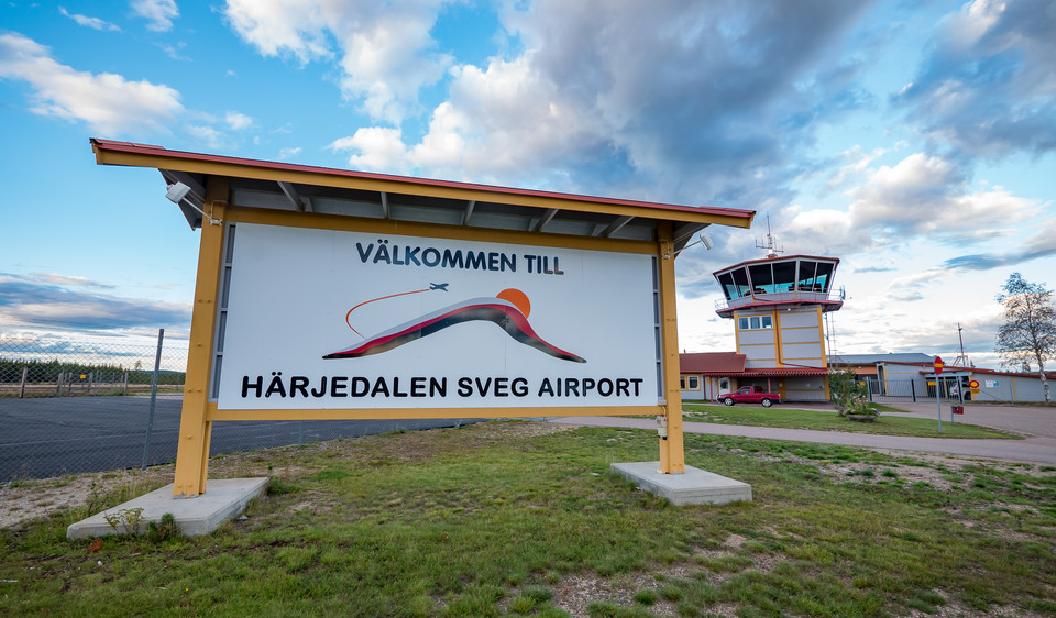 Skylt på gräsmatta med texten Välkommen till Härjedalen Sveg airport framför ett staket och en byggnad med ett trafiktorn.
