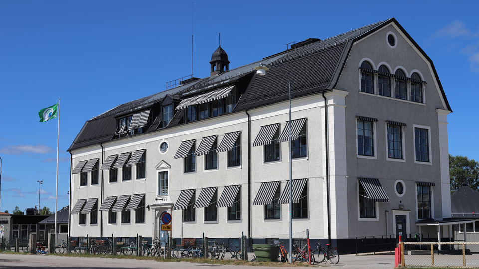 Grå äldre rappad trevåningsbyggnad med svart tak mot en klarblå himmel.