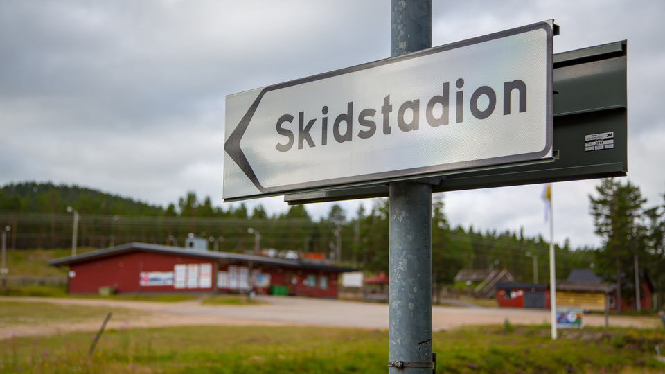 Vägskylt som pekar mot Skidstadion med gröna gräsmattor, grusplan och ett rött hus.