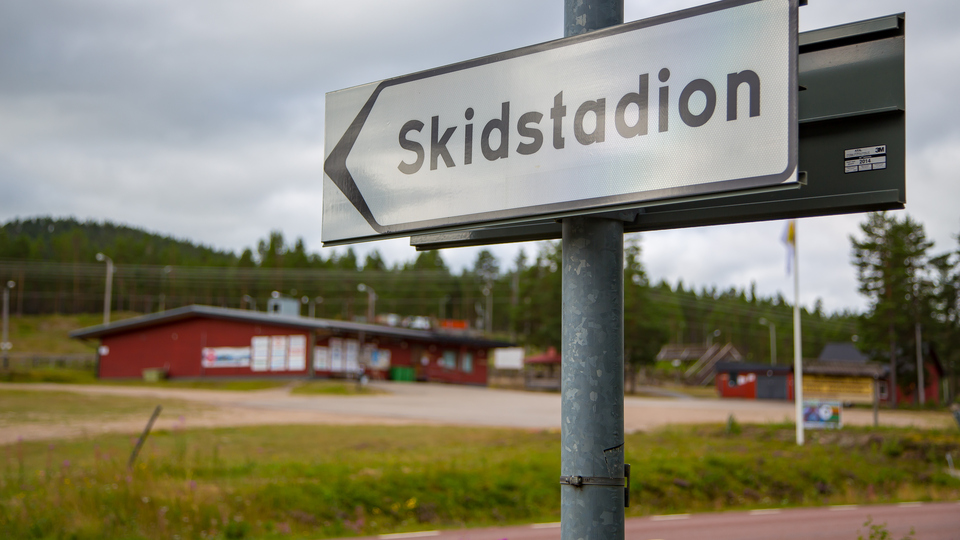 Vägskylt som pekar mot Skidstadion med gröna gräsmattor, grusplan och ett rött hus.