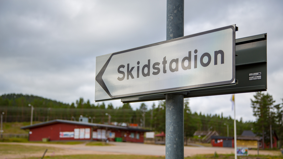 Vägskylt som pekar mot Skidstadion med gröna gräsmattor, grusplan och ett rött hus.