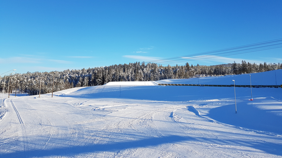 Snötäckt yta med en skidskyttevall i bakgrunden. Klarblå himmel och granskog i fonden.