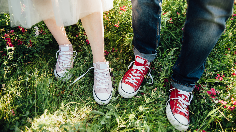 Två par fötter i rosa och röda lätta sommarskor med knytning står i grönt gräs. En person är barbent och har en flortunn kjol, en person har blå jeans som är uppvikta nertill.