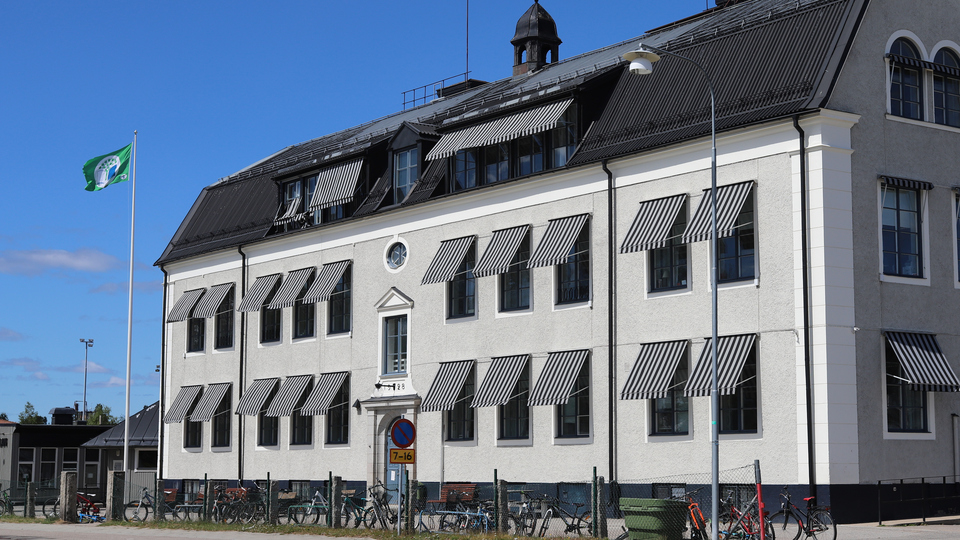 Grå äldre rappad  trevåningsbyggnad med svart tak mot en klarblå himmel.