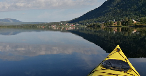 Fören på en kanot  på en spegelblank sjö med en by och ett berg i fonden.