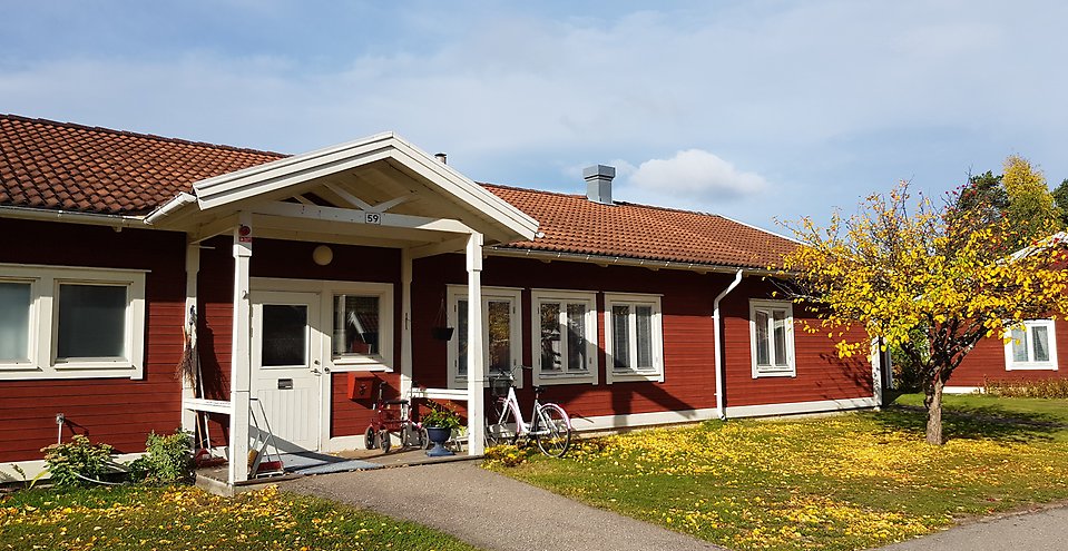Rött enplanshus med vita detaljer. Vid entrén en cykel och en rollator. Framför huset ett träd med höstfärger. 