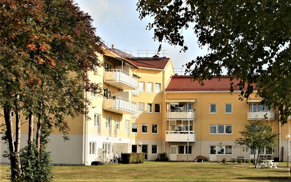 Gult flervåningshus med vita balkonger och träd i förgrunden.